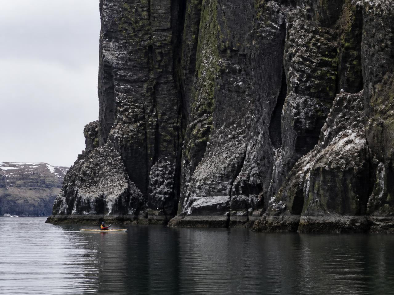 A lone sea kayaker paddling beneath towering basalt sea cliffs